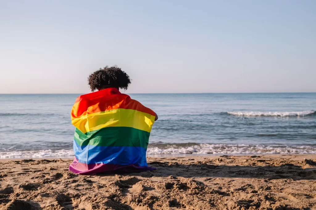 person-wrapped-in-rainbow-flag-sits-on-beach